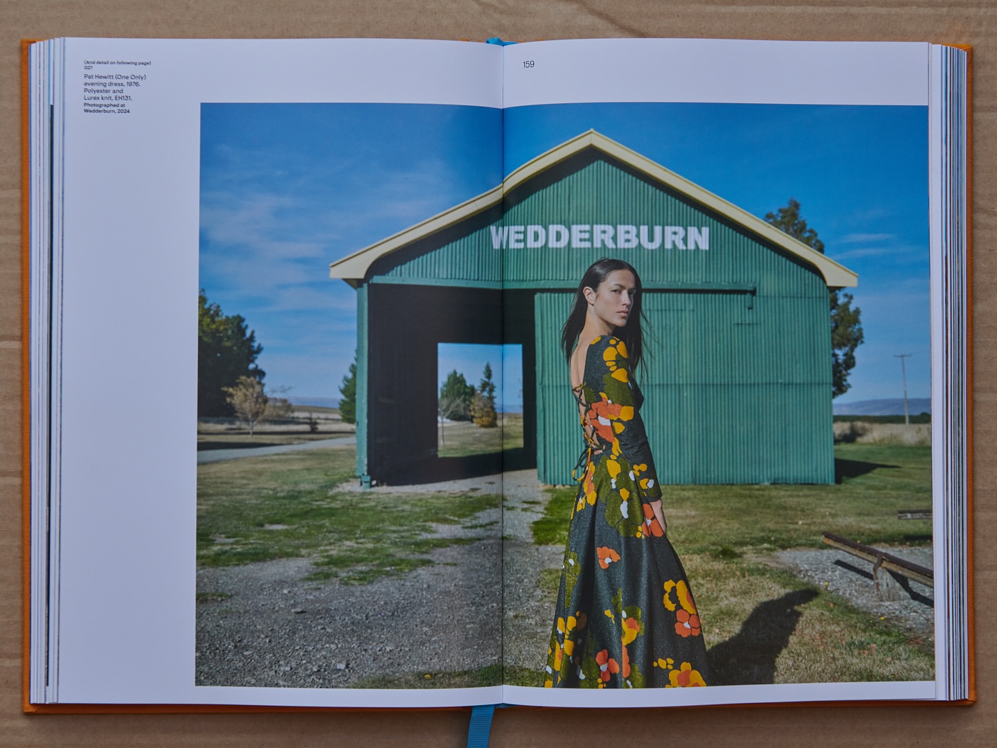 Woman in floral gown stands before a green storage shed with WEDDERBURN painted on its face, set against a blue sky and grassy field.