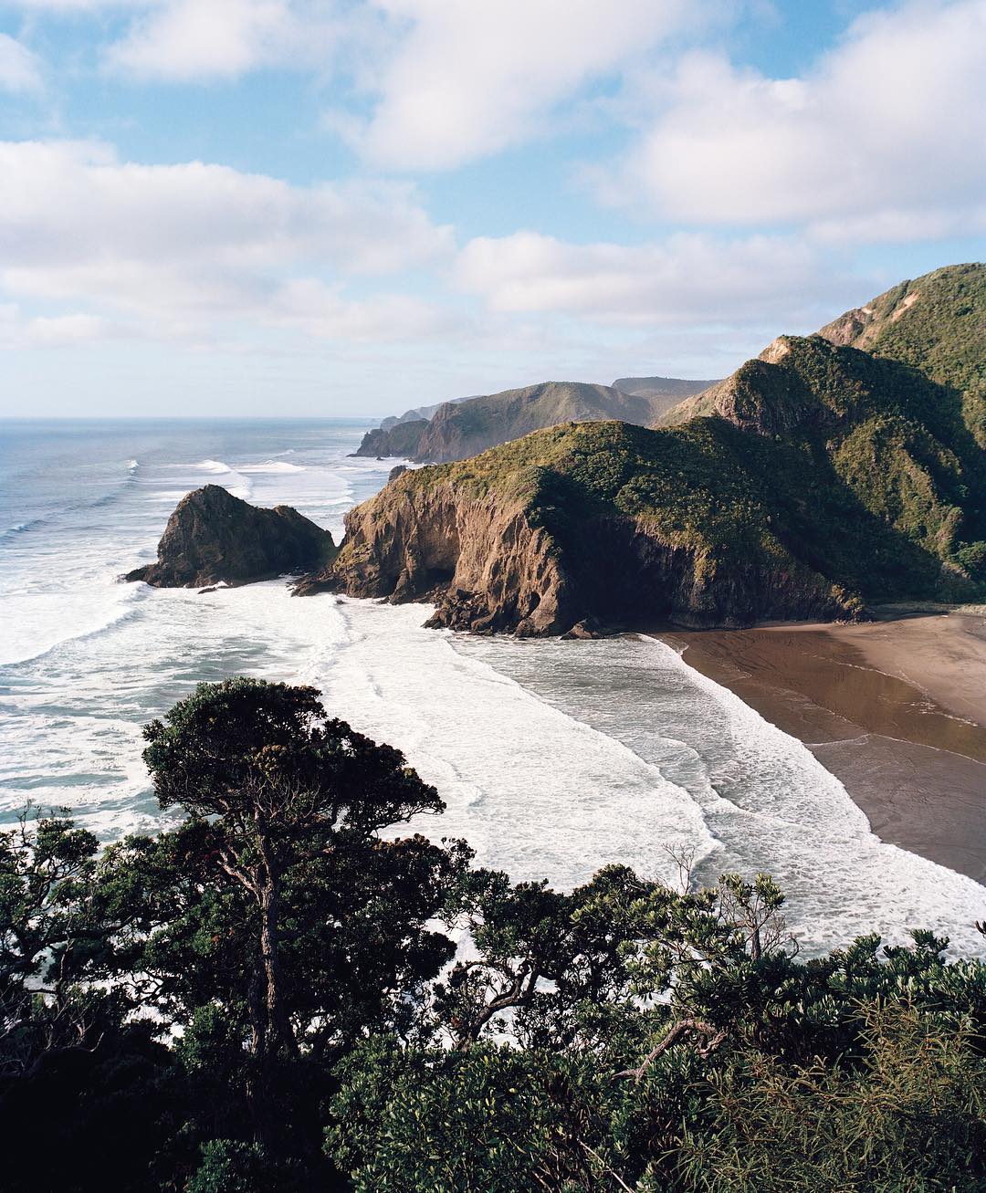 Scenic view of a beach with dark sand and white waves, framed by a lush green hillside under a blue sky with scattered clouds.