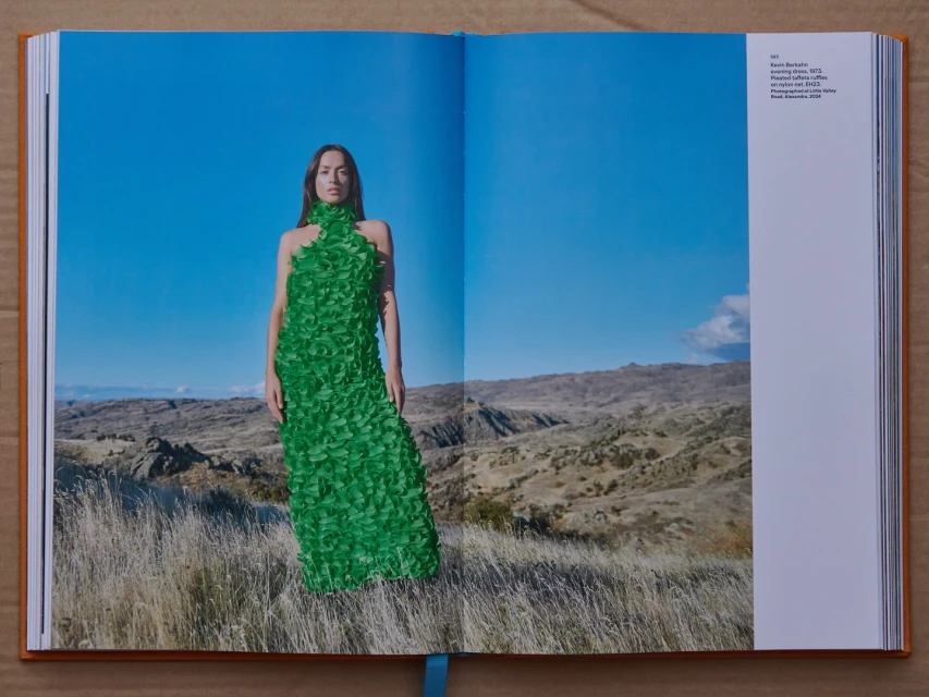 A fashion shot in an open book features a woman in a green dress standing in a grassy field with a mountain range and blue sky behind.