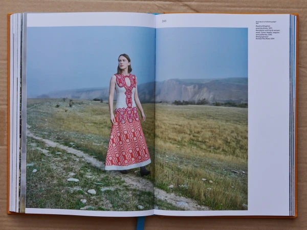 Model standing on a dirt path wearing an off-white evening dress with intricate red embroidery, with rolling hills in the background.