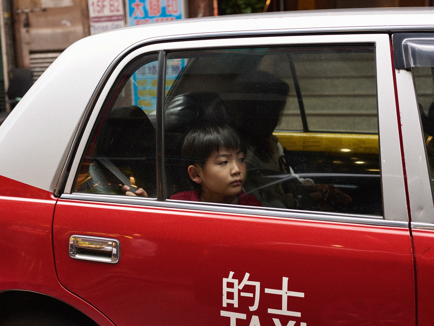 Young boy gazes out the window of a red and white taxi, with urban buildings blurred in the background, creating a candid moment.