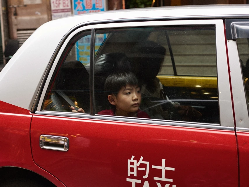 Young boy gazes out the window of a red and white taxi, with urban buildings blurred in the background, creating a candid moment.