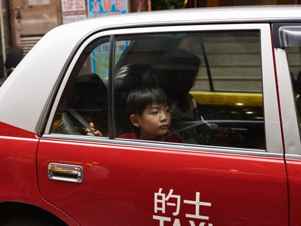 Young boy gazes out the window of a red and white taxi, with urban buildings blurred in the background, creating a candid moment.