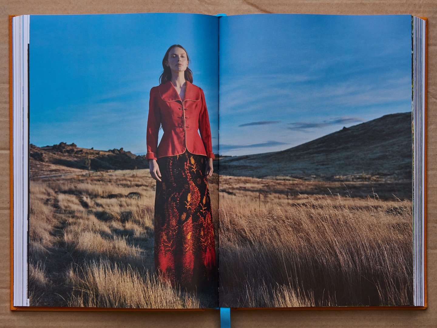 Model in a red jacket and patterned skirt standing in a dry grassy landscape under a blue sky photographed by Derek Henderson.