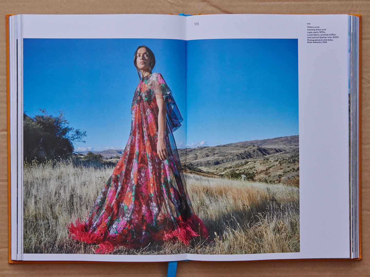 An open book displaying a fashion editorial photograph of a woman in a floral maxi dress with feather trim standing in a dry field.