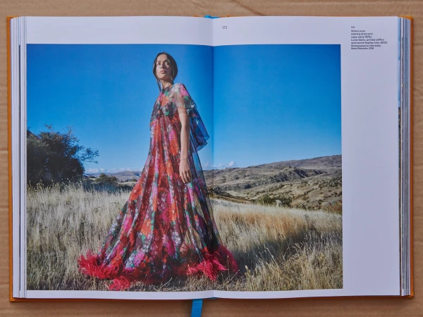 An open book displaying a fashion editorial photograph of a woman in a floral maxi dress with feather trim standing in a dry field.
