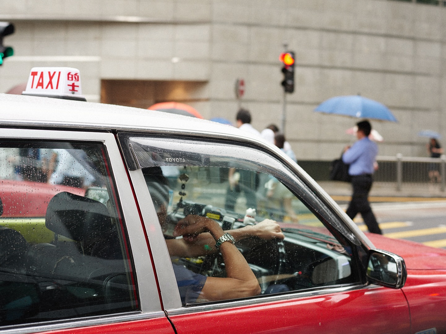 Hong Kong taxi cab parked on a city street, reflecting light and passersby in its windows; pedestrians and buildings in background.