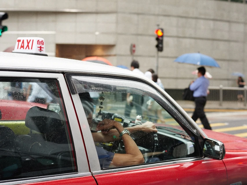 Hong Kong taxi cab parked on a city street, reflecting light and passersby in its windows; pedestrians and buildings in background.