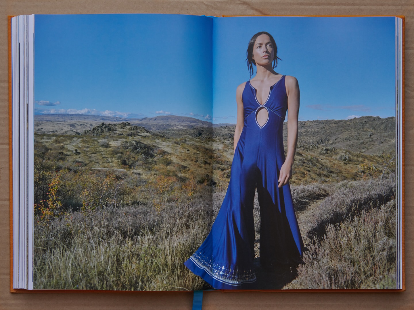 Fashion shot from an open book features a woman wearing a blue jumpsuit standing in a grassy field against a clear blue sky and mountain range.