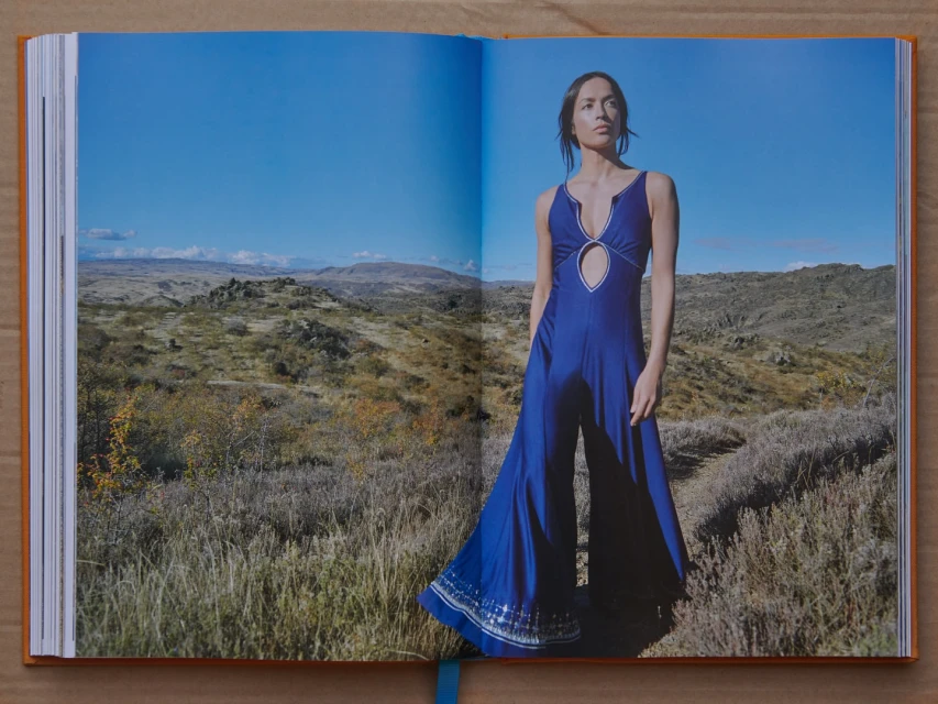 Fashion shot from an open book features a woman wearing a blue jumpsuit standing in a grassy field against a clear blue sky and mountain range.