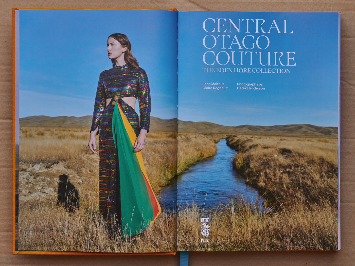 Open book featuring a fashion photograph of a model in a colorful dress in a field and a landscape with a river and mountains.