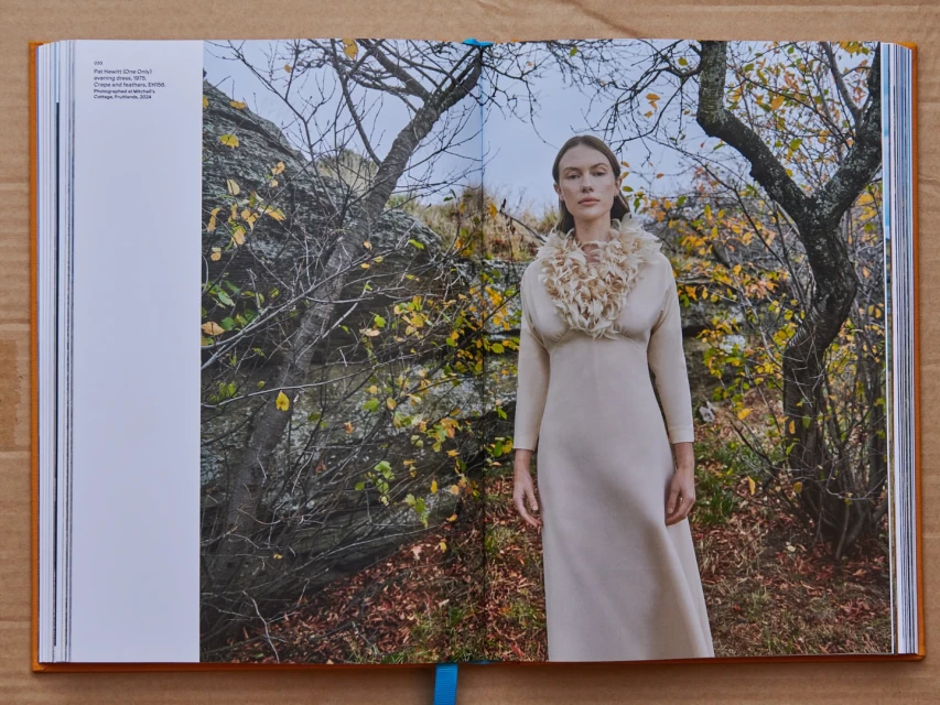 Fashion editorial image featuring a woman in a long dress with a feathered collar, posing outdoors in a natural setting with trees and rocks.