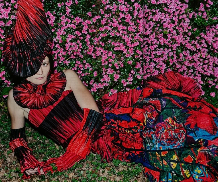 Fashionable individual in vibrant pleated red and black ensemble lies amidst lush greenery and flowering pink azalea bushes.
