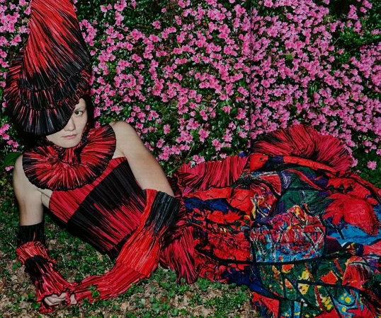 Fashionable individual in vibrant pleated red and black ensemble lies amidst lush greenery and flowering pink azalea bushes.