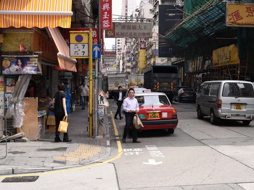 Busy Hong Kong street scene features pedestrians on the sidewalk and taxis, vans, and trucks moving along the narrow roadway.