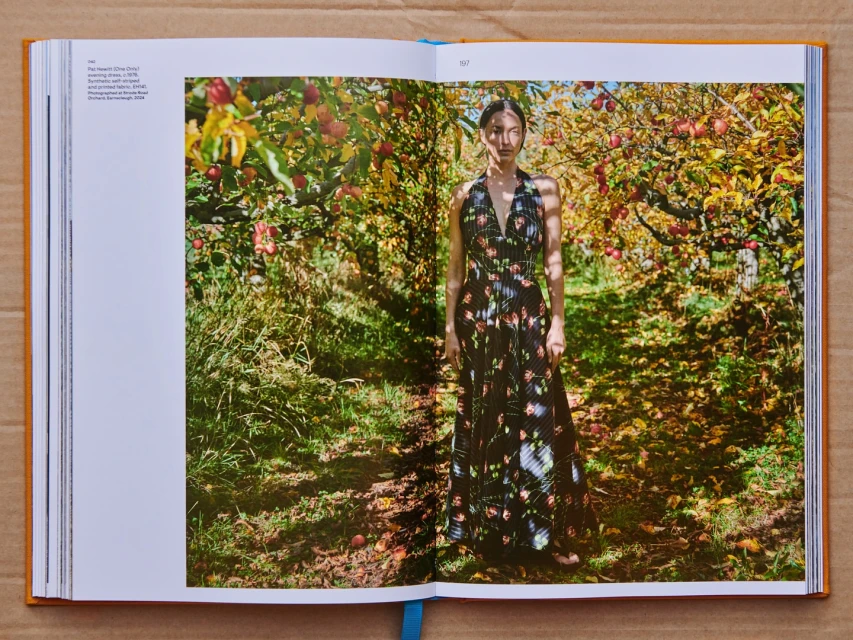 Fashion editorial photo of a young woman in a floral dress standing in an apple orchard with green grass and fallen leaves.