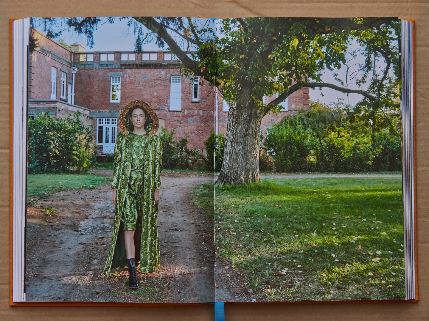 Fashion model in green patterned outfit poses on a dirt path in front of a red brick building with large tree and green lawn.