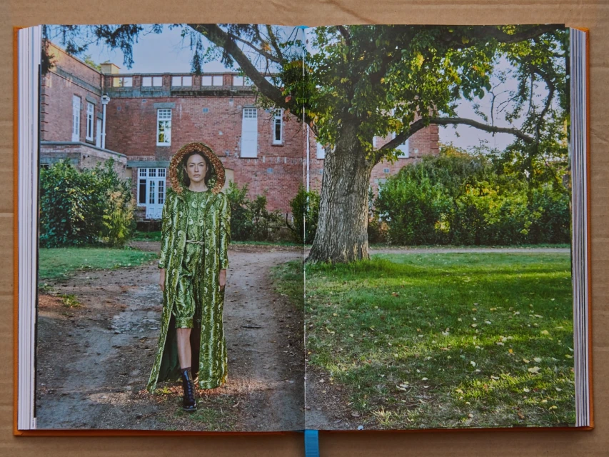 Fashion model in green patterned outfit poses on a dirt path in front of a red brick building with large tree and green lawn.