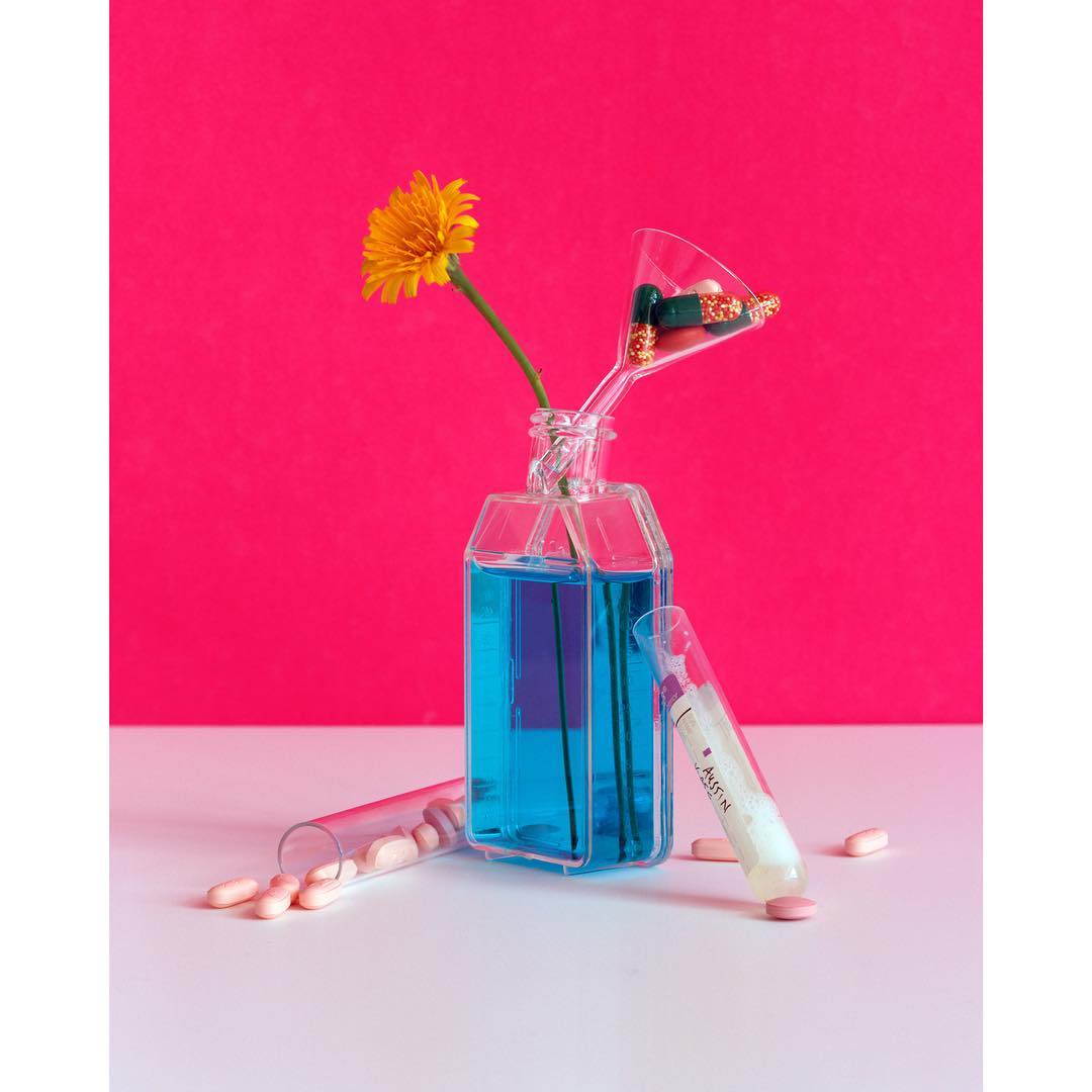 Still life photo of a blue liquid in a clear container with a yellow flower, capsules in a glass, and pills on a white surface.