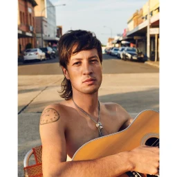 Shirtless man with a mullet hairstyle and shoulder tattoo holding an acoustic guitar on a sunny street by Derek Henderson.
