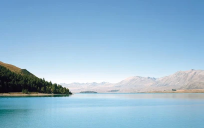 Wide scenic view of a calm turquoise lake bordered by a forested green hill on the left and distant dry mountain ranges under a clear blue sky.
