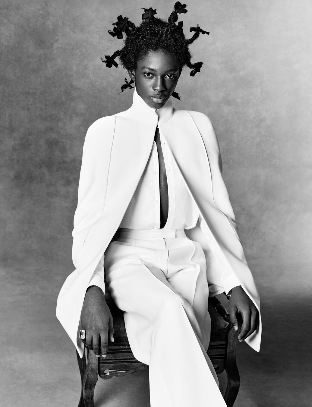 Portrait of an African American adult female model with a modern hairstyle sitting on a wooden chair in a monochromatic shot.