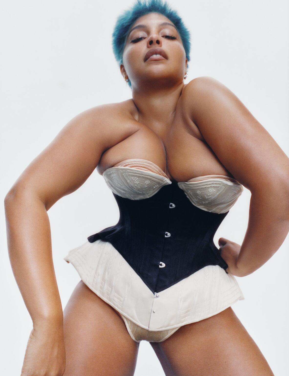 A woman with blue hair is modeling a black corset with a beige bra underneath, against a white backdrop, in a medium-angle studio shot.