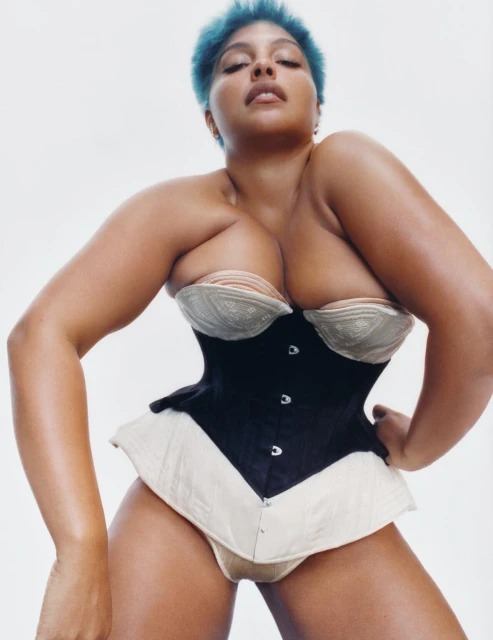 A woman with blue hair is modeling a black corset with a beige bra underneath, against a white backdrop, in a medium-angle studio shot.