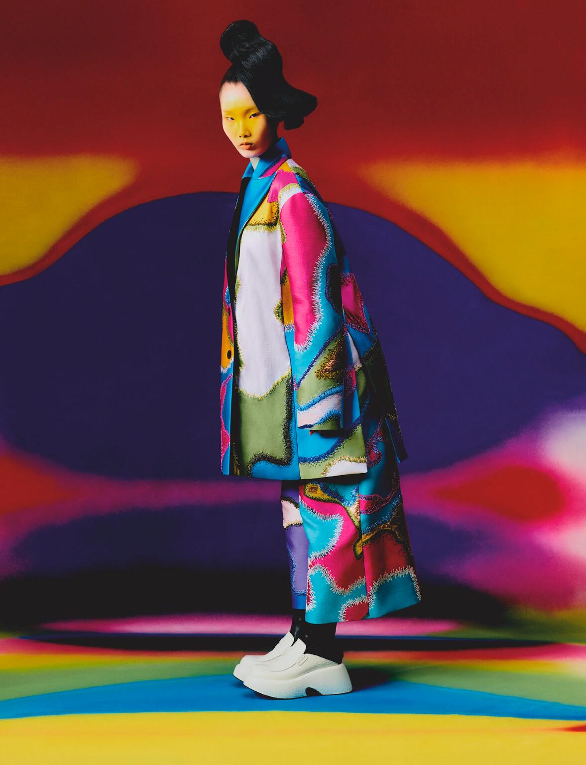 Woman wearing a vibrant, patterned coat and platform shoes poses in front of a colorful, abstract backdrop for a fashion editorial.