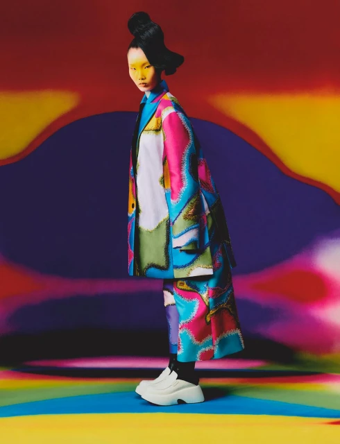 Woman wearing a vibrant, patterned coat and platform shoes poses in front of a colorful, abstract backdrop for a fashion editorial.