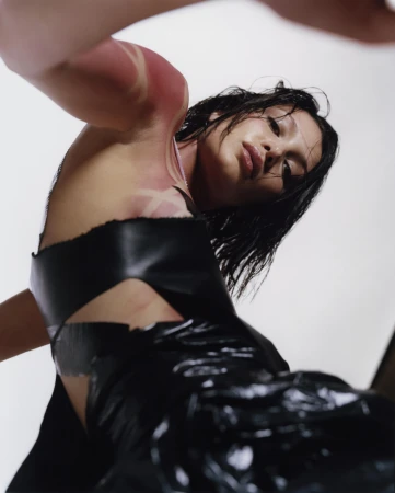 Adult female with wet black hair in dark avant-garde fashion dress, shot at a low angle in a photo studio against a white background.