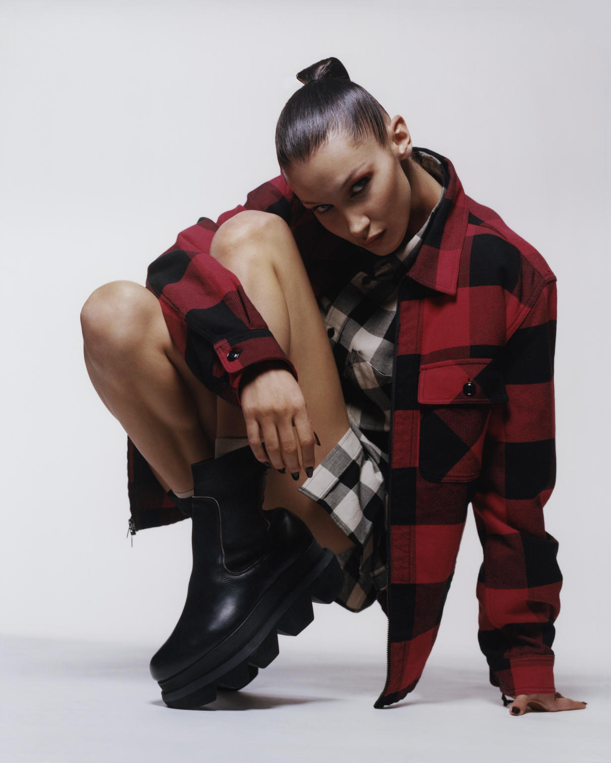 Portrait of a model in a red and black plaid jacket and black boots squatting in a photo studio against a white backdrop.