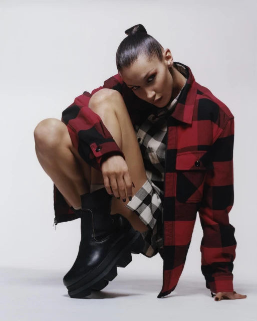 Portrait of a model in a red and black plaid jacket and black boots squatting in a photo studio against a white backdrop.