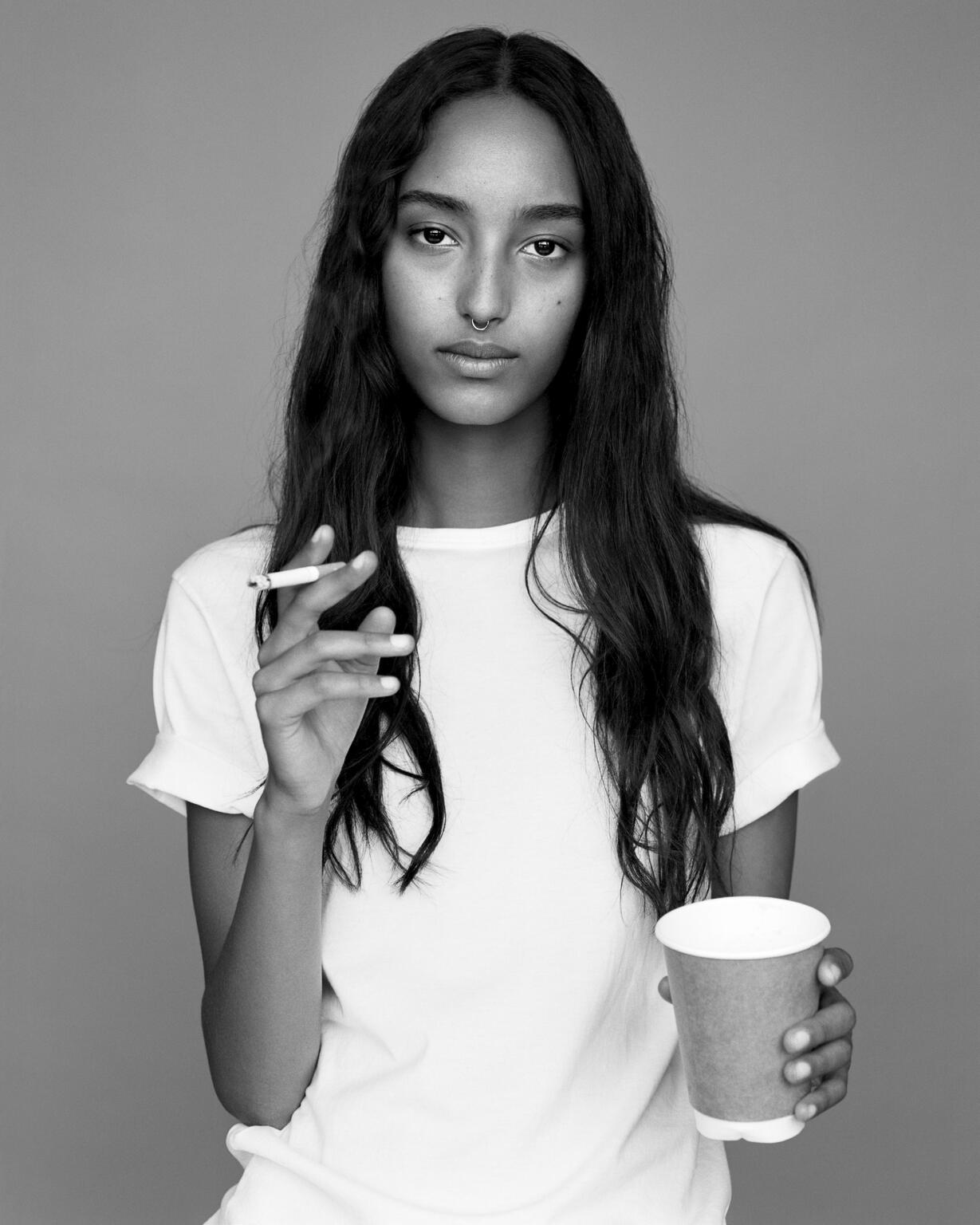 Black and white portrait of a slender young woman with long dark hair holding a cup and a cigarette indoors on a grey background.