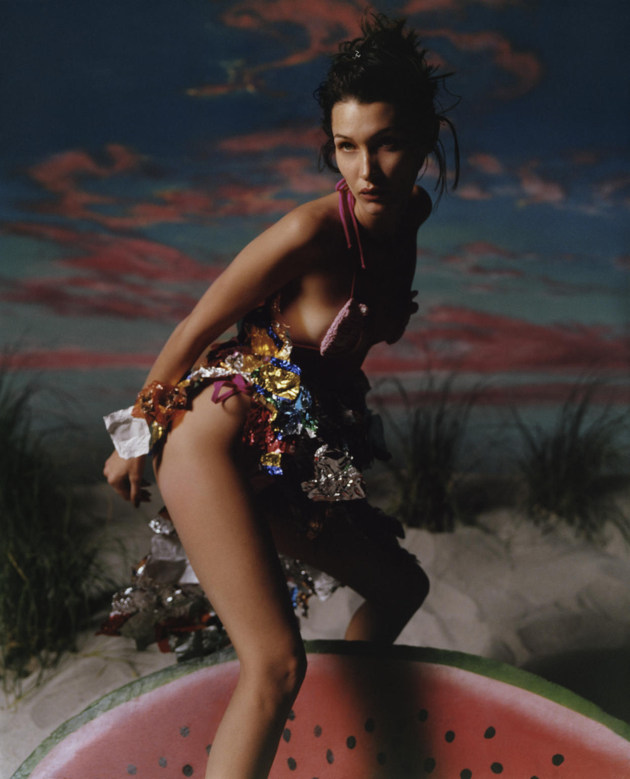 Woman in a creative outfit posing on a watermelon slice on a sandy beach with an expressive sky; fashion, beauty, portrait photography.