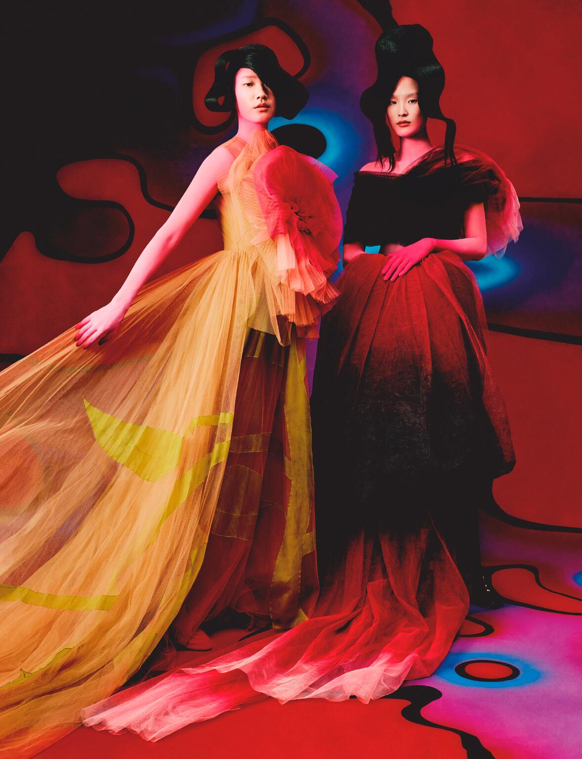 Two Asian women pose in high-fashion tulle gowns in a studio with a red, black, and blue abstract background with a light blue lit floor.