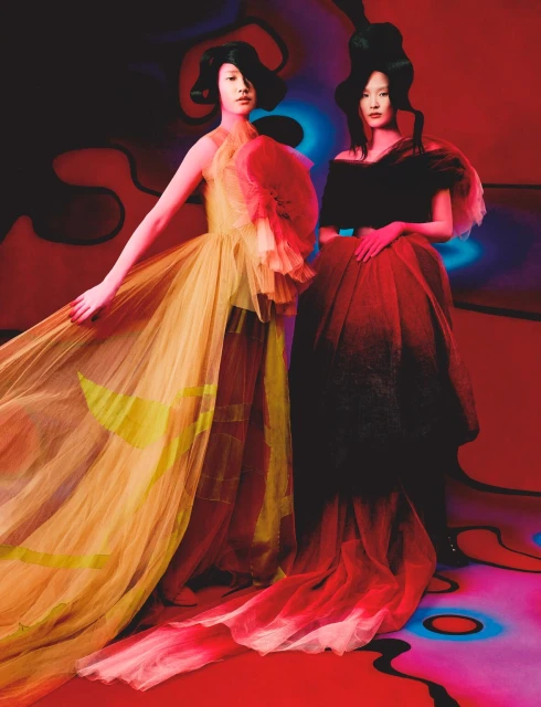 Two Asian women pose in high-fashion tulle gowns in a studio with a red, black, and blue abstract background with a light blue lit floor.