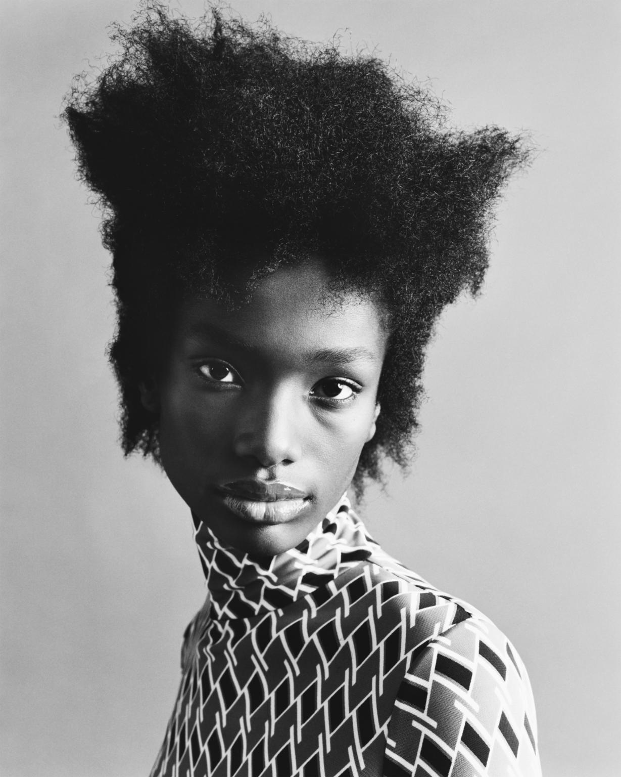 Striking portrait of a Black female model with an afro, wearing a geometric patterned top, against a neutral grey backdrop.