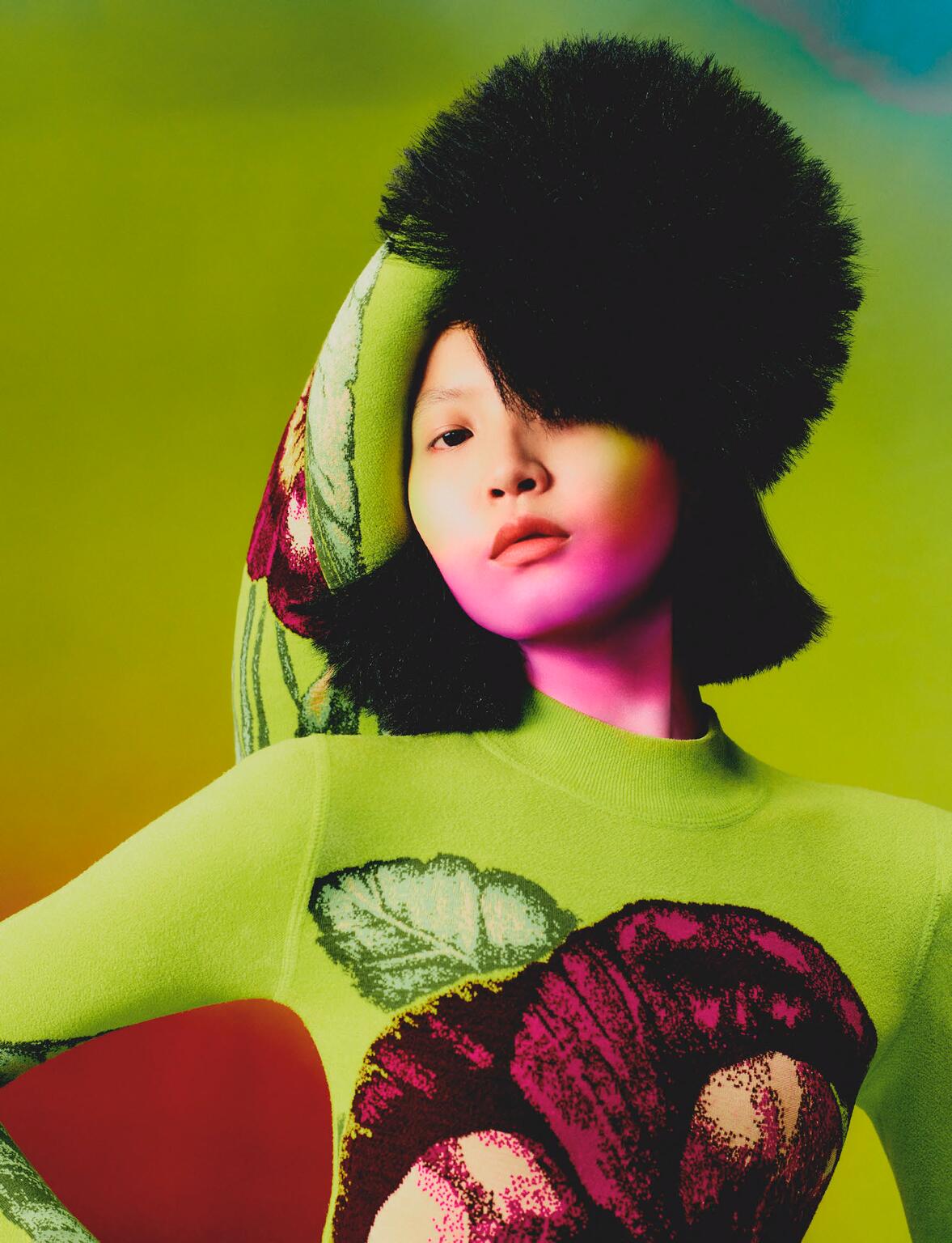 Portrait of a female model in a neon green flower-print dress and furry hat, posing against a vibrant, colorful gradient background.
