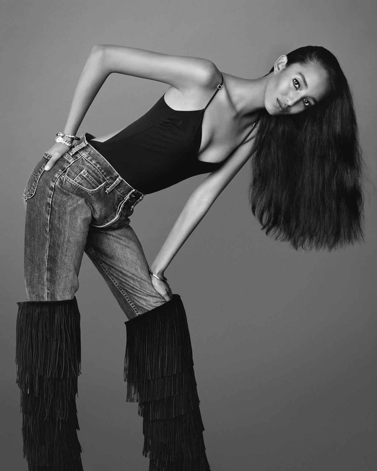 A woman with long hair poses in a black bodysuit and jeans with fringed leg coverings in a studio against a gray backdrop.