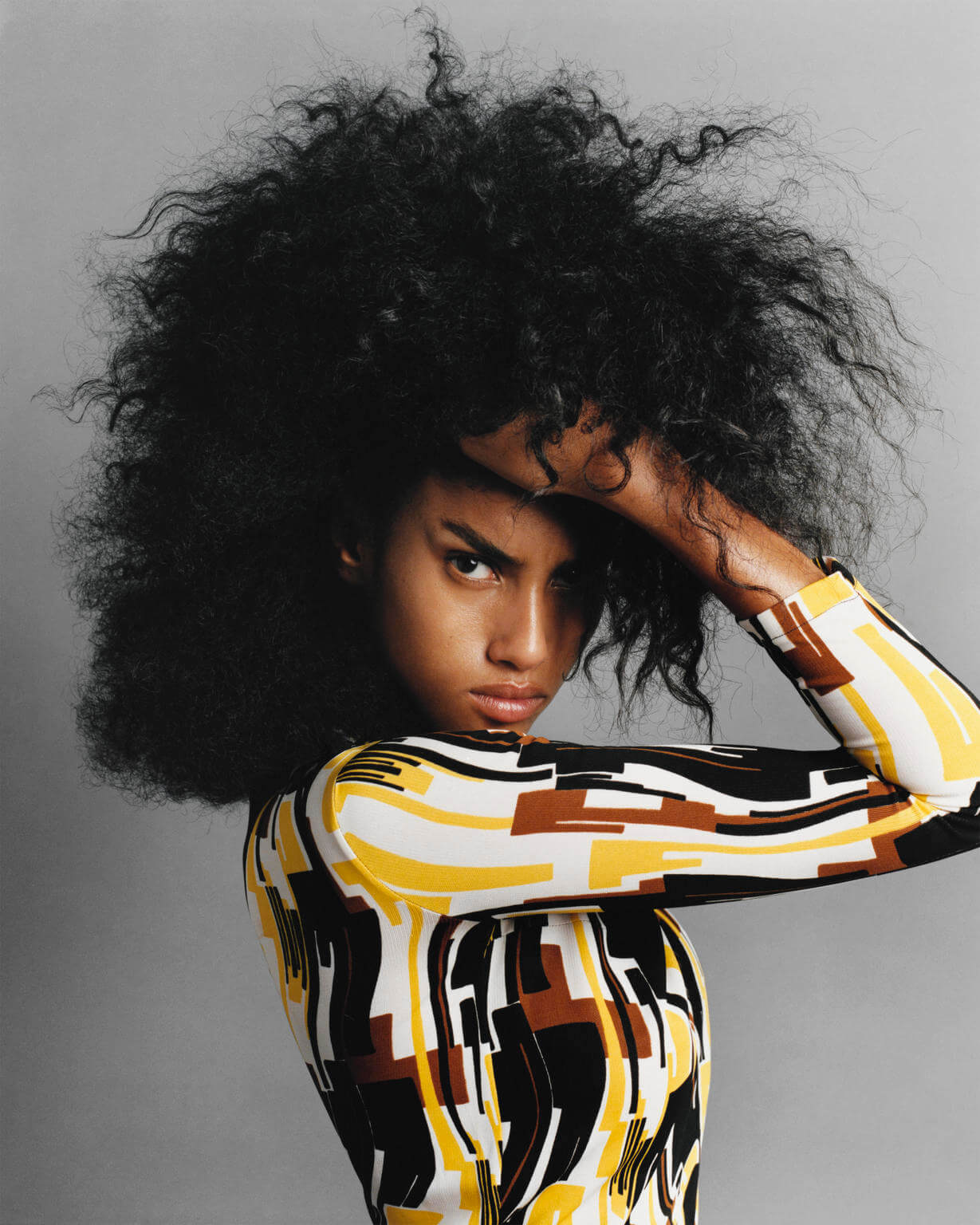A fashionable person with striking afro-textured hair poses in a studio, wearing a modern geometric patterned long-sleeve top and makeup.