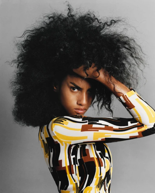 A fashionable person with striking afro-textured hair poses in a studio, wearing a modern geometric patterned long-sleeve top and makeup.