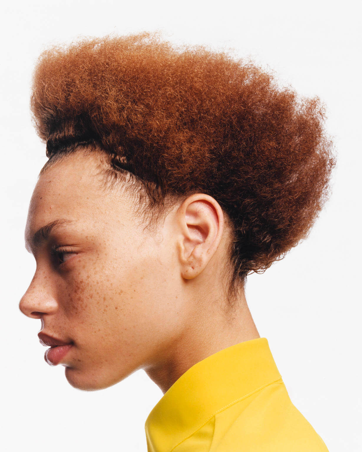 Profile portrait of adult female with short brown hair, freckled skin, and yellow top against a white backdrop in a photo studio.