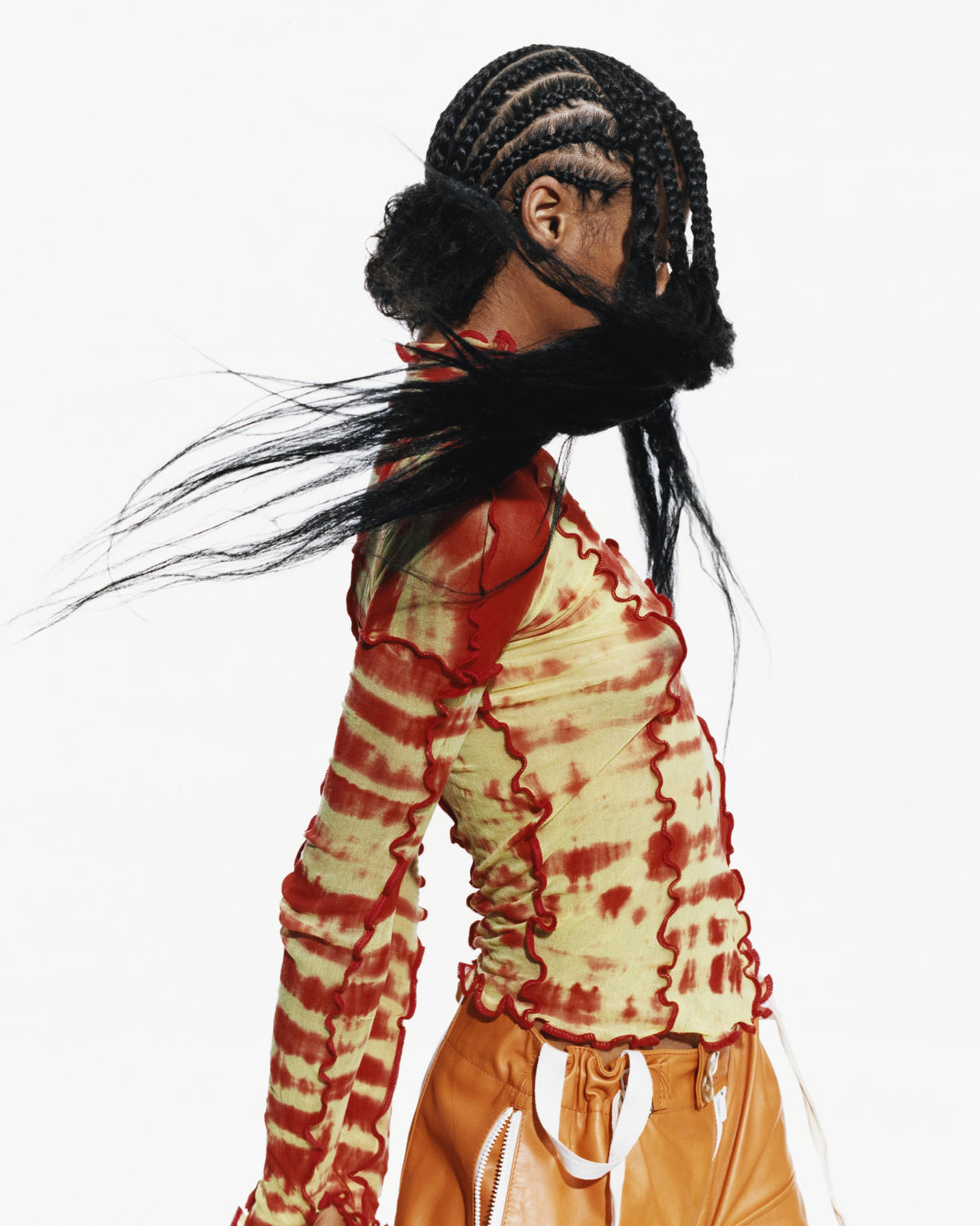 A person with braided hair wearing a yellow and red long sleeve shirt, and orange pants poses against a white backdrop in studio.