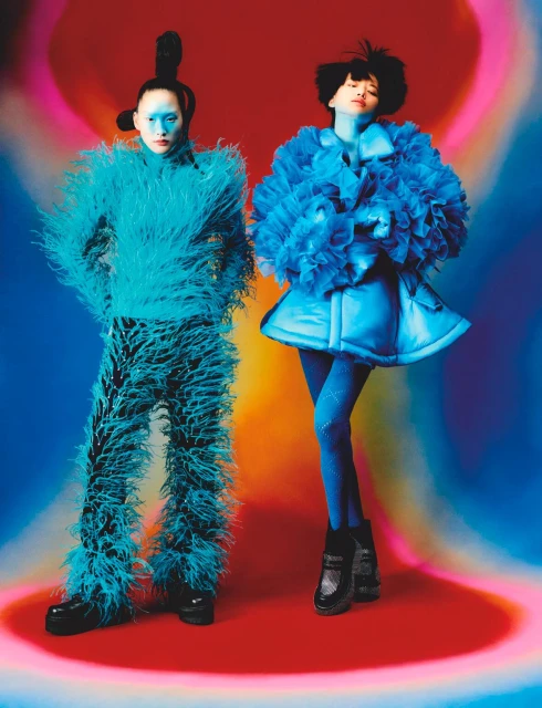 Two models in futuristic fashion pose against a vibrant, swirling backdrop. One wears a feathery blue suit, the other a ruffled dress.