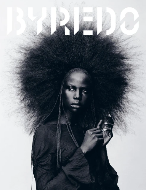 Striking black and white portrait of a woman with a voluminous afro hairstyle, wearing a dark top, against a light backdrop. 