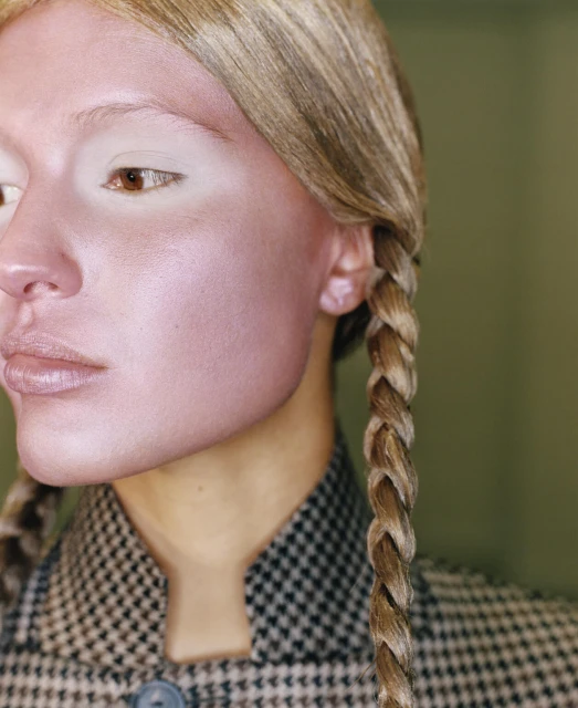 Close-up of a woman with blonde braided pigtails and light skin. She wears a houndstooth jacket, with a soft green background.