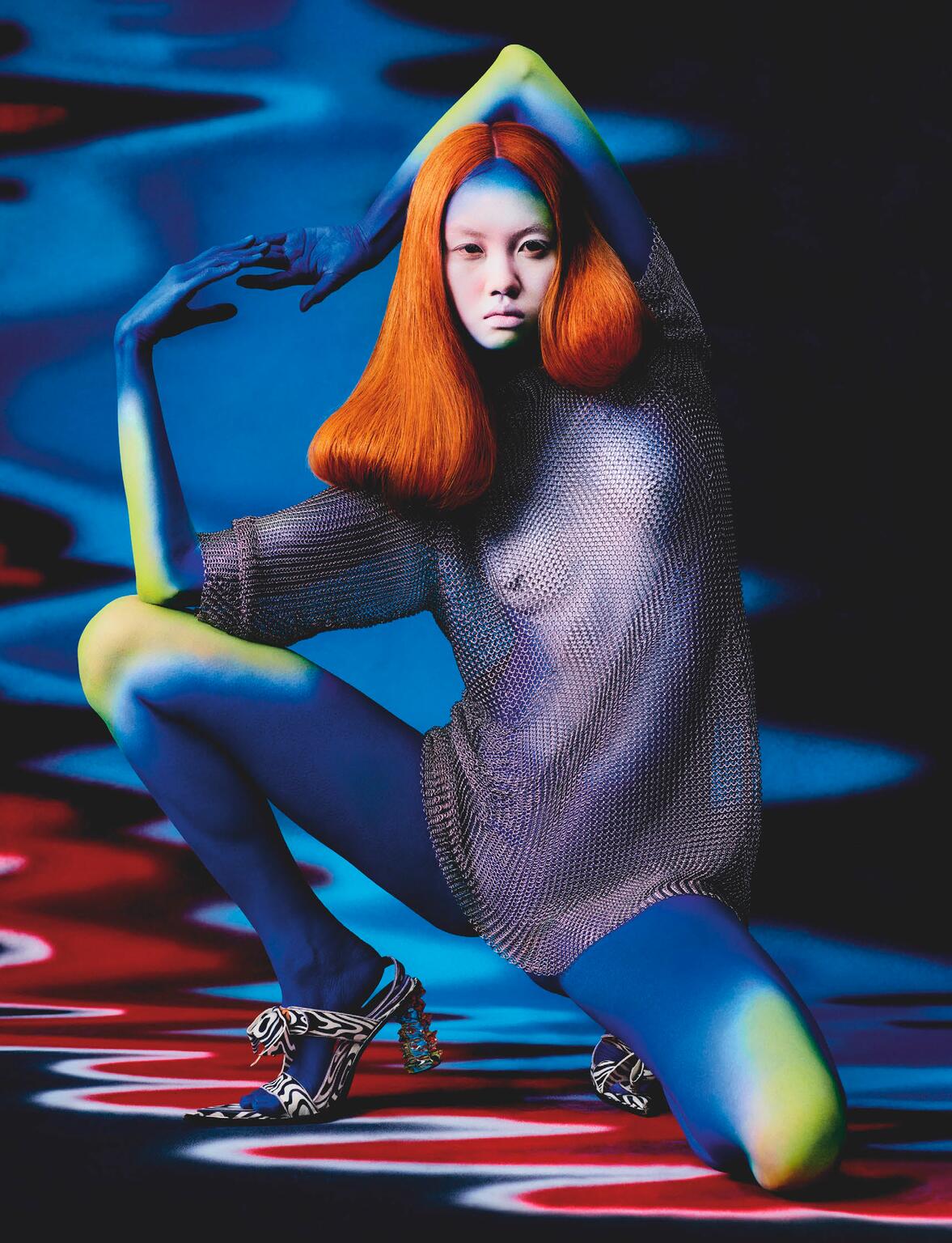 Person with orange bob hairstyle and blue painted skin poses in a silver metallic knit top and zebra-print high-heeled sandals.