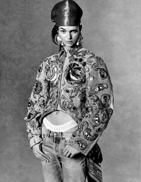 Monochrome portrait of a woman wearing an ornately embellished jacket, jeans, large earrings, and a head wrap in a studio setting.