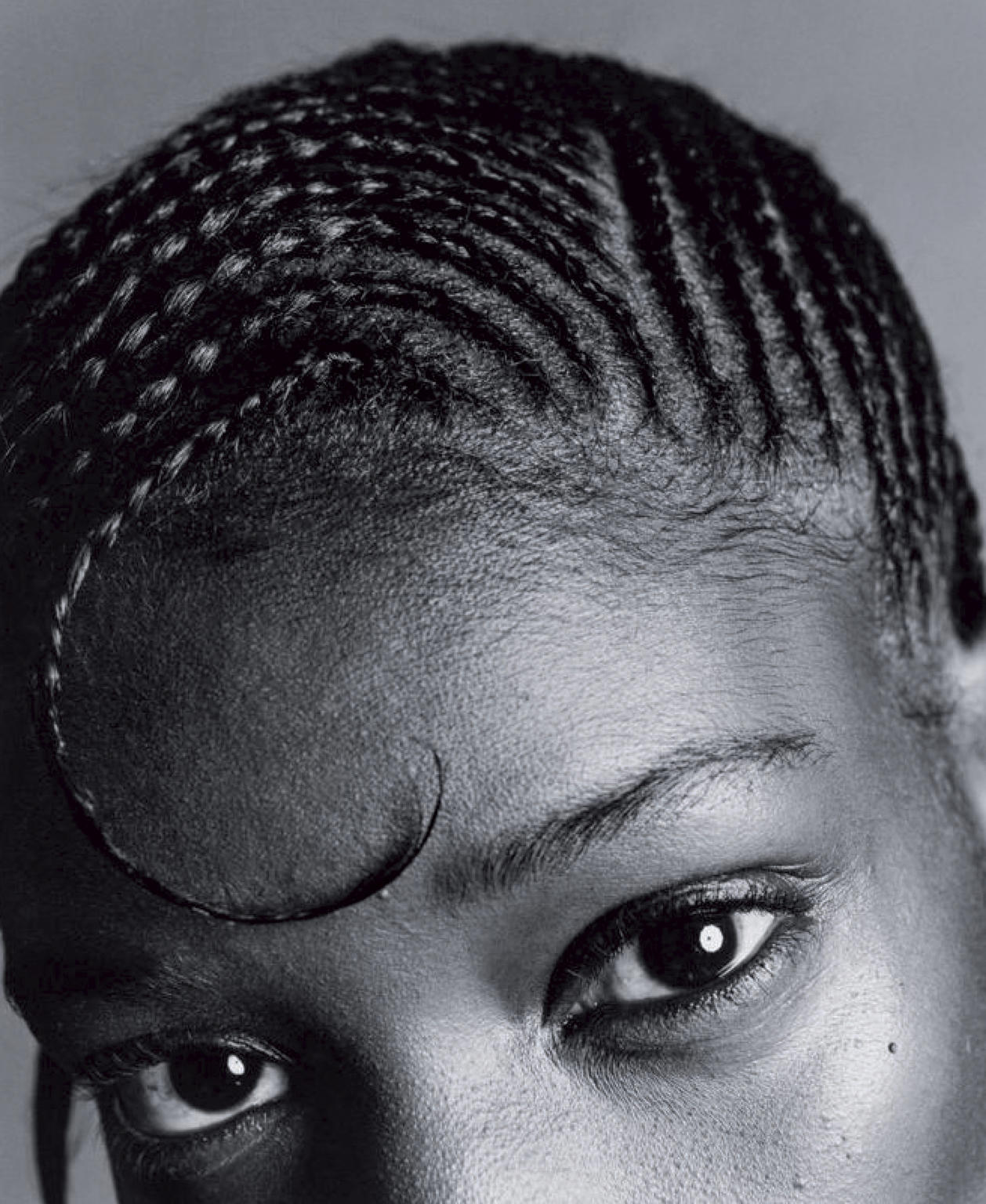 Close-up studio shot of a Black adult female with braided hair and distinct swooped forelock, eyes looking directly at the camera in monochrome.